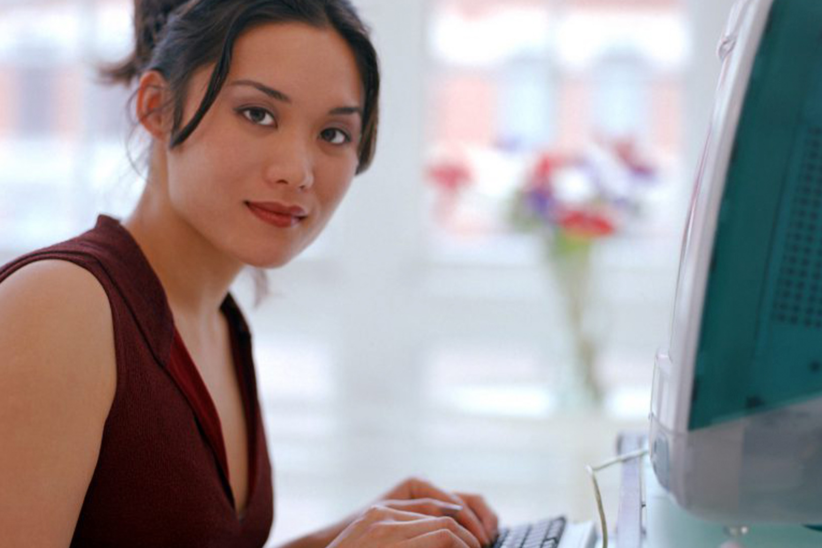 A woman smiling slightly and looking towards the camera while her hands rest on the keyboard of an old Mac desktop computer.