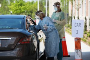 Two health care workers in scrubs, plastic protective gear and masks speak with a person through the driver's window of a vehicle. 