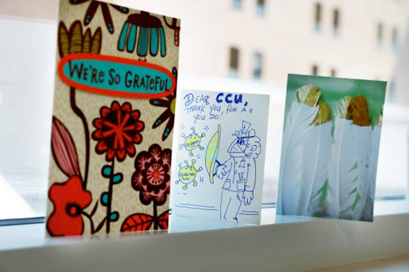 Three hand-made cards wishing patients of coronavirus well, with text We’re So Grateful, flower designs, cartoon drawings and feathers on a windowsill.