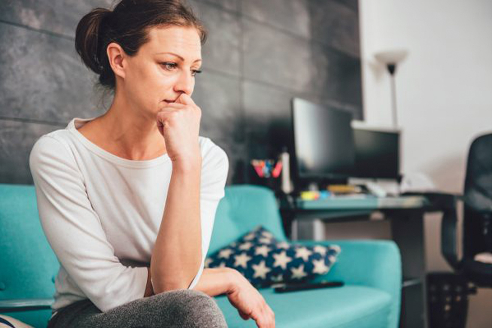 A woman looks worried as she sits on a teal couch, legs crossed, hand to her mouth.