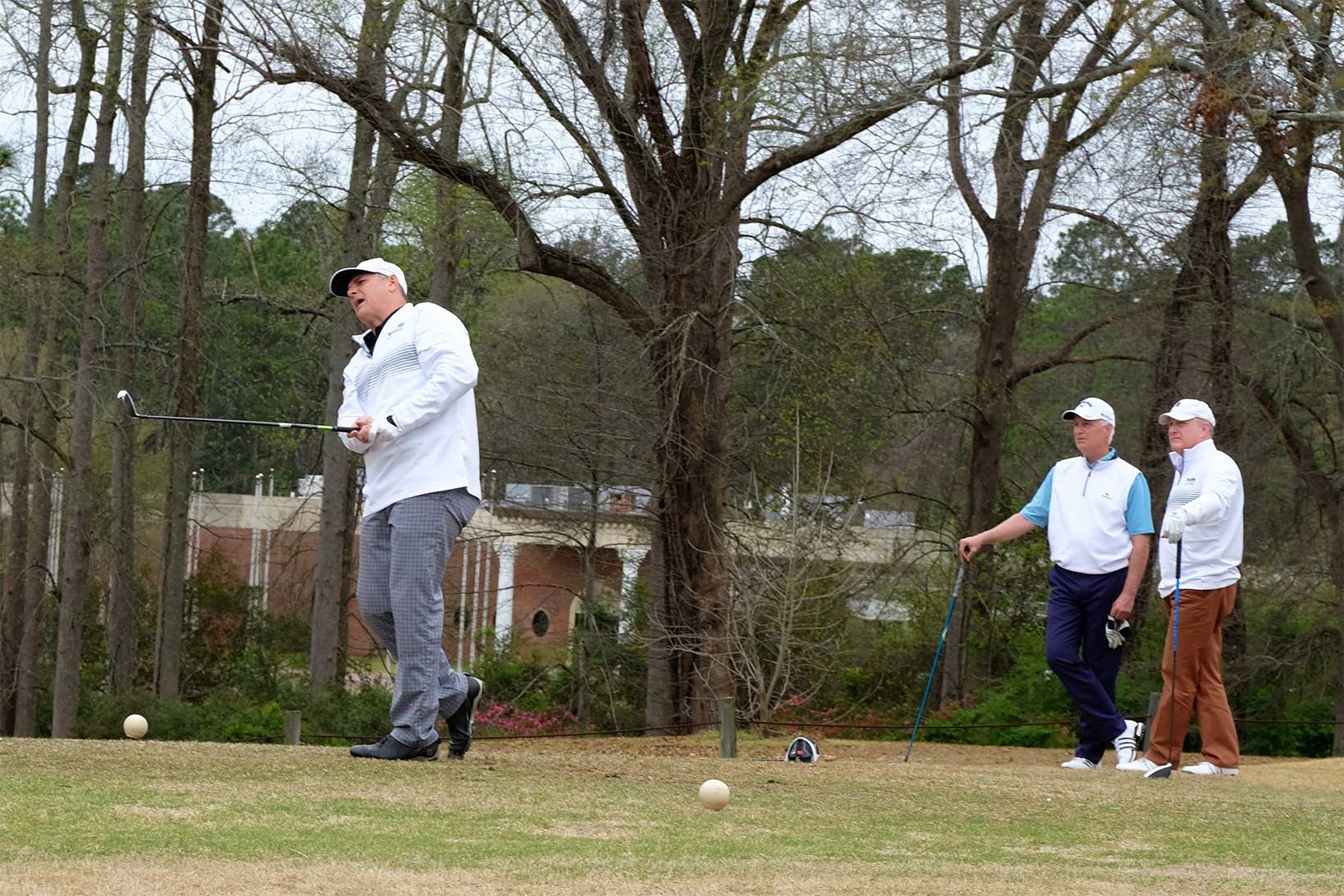 Three men playing golf, with two in the background observing the shot of another man, still in his golf stance, grimacing at his performance.