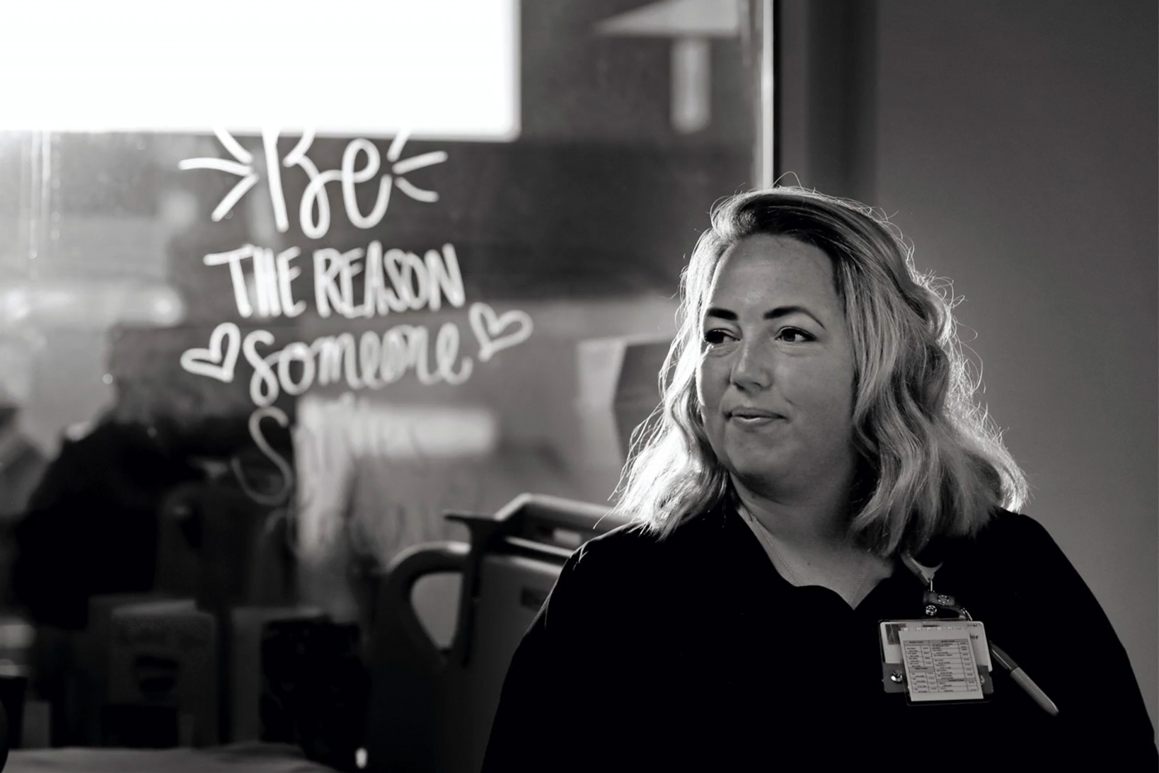 Black and white photo of Theresa Kossler, smiling in front of a window with calligraphy reading Be the reason someone smiles today.