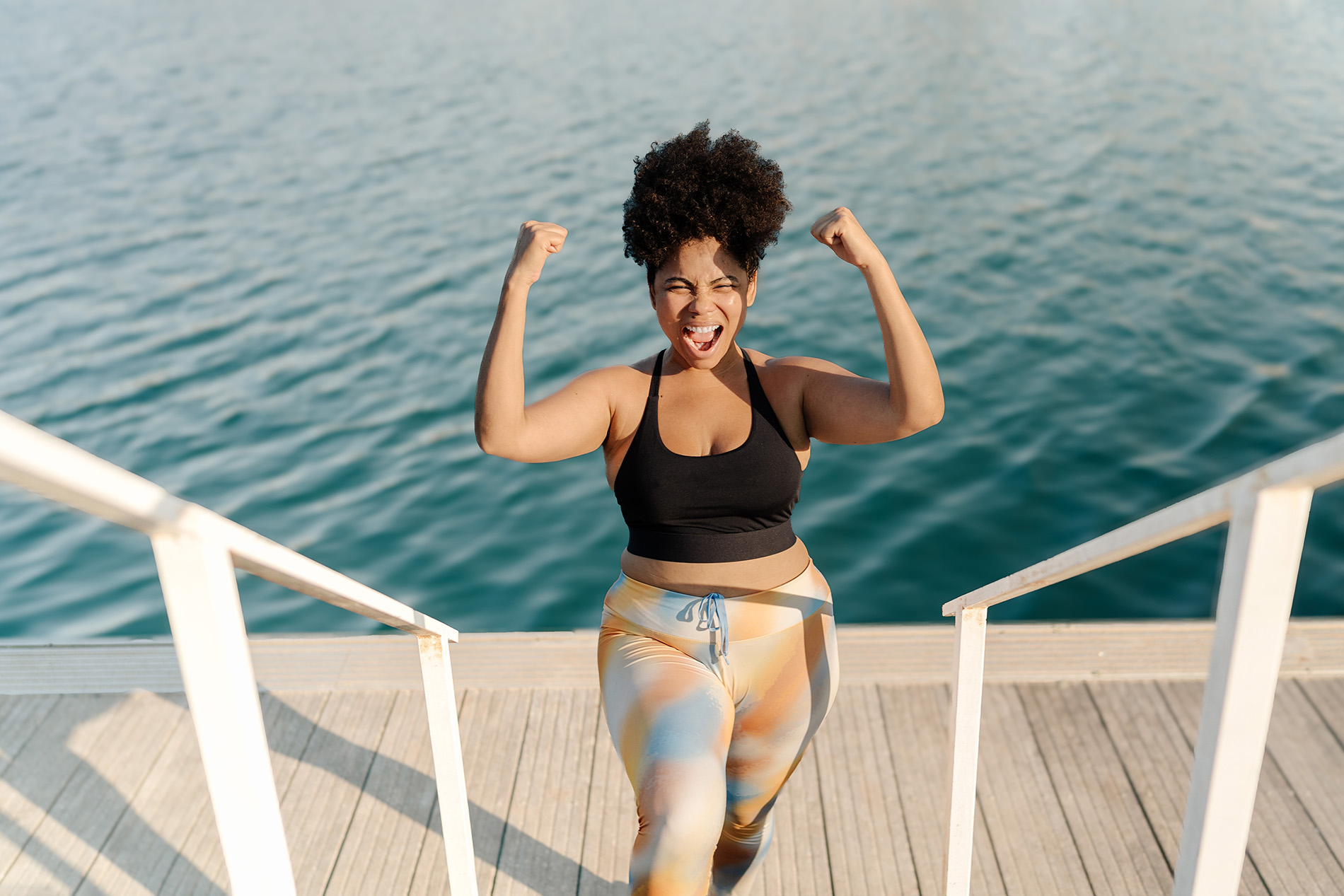 overweight black woman holding hands up in celebration on a dock by a lake