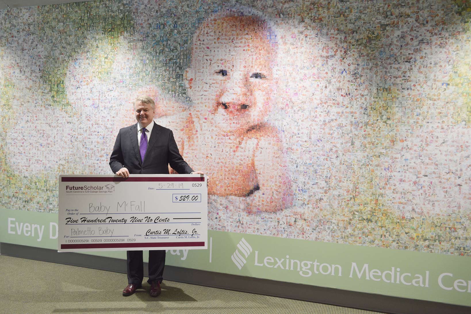 Special Delivery – A Palmetto Baby 529 Plan Grant Treasurer Loftis holding a large check addressed to Baby McFall posing in front of the baby mosaic in the new patient care tower.