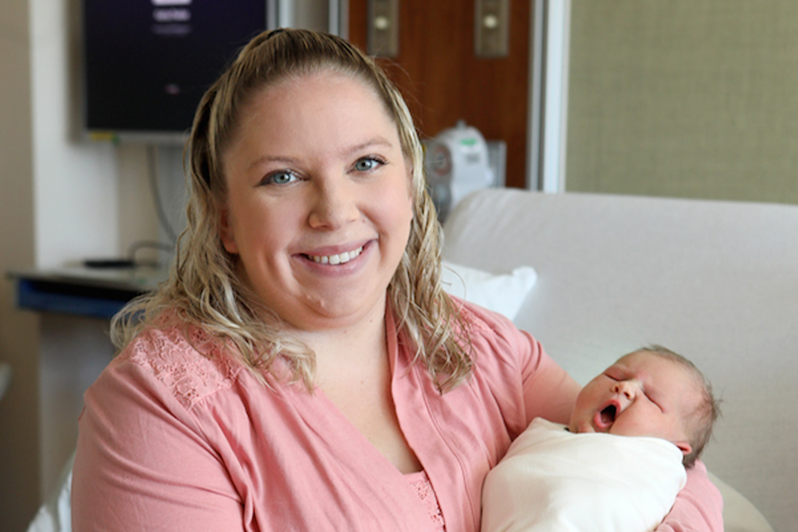 A portrait of Katlyn in a pink hospital gown, sitting on the hospital bed and smiling while holding her newborn baby, Savannah Kate, who is yawning.