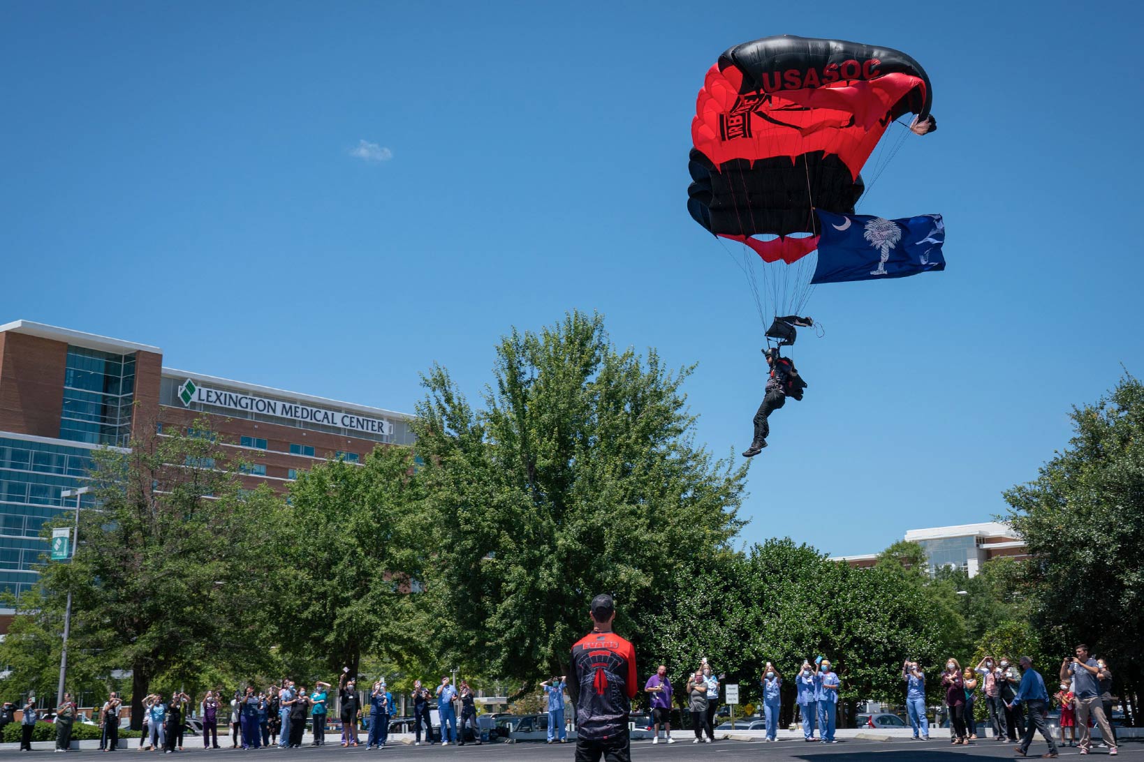 A Black Dagger member parachuting on the Lexington Medical Center main campus, waving the South Carolina flag, as staff and patients watch.