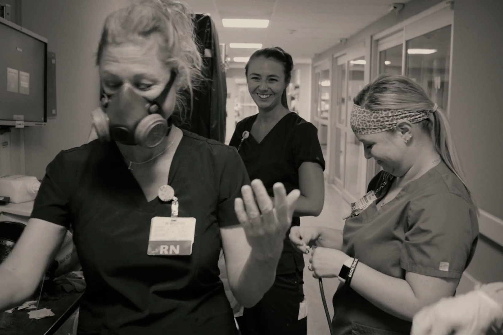 Black and white shot of nurse Kim Johnson in a safety respiratory mask joking with two colleagues, all in scrubs.